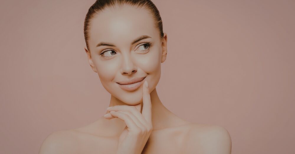 A woman with a smooth, lifted facial profile resting her finger on her chin against a soft pink background