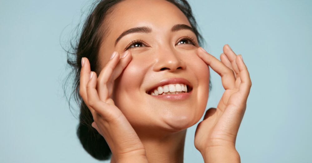 Smiling woman gently touching her face with both hands against a light blue background showing smooth healthy skin