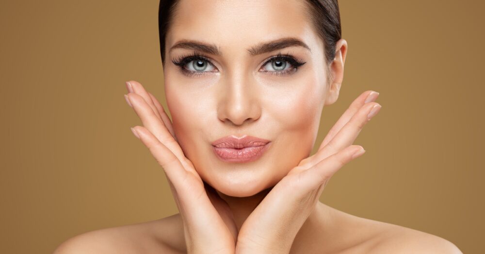 Close up of a woman gently cradling her face with her hands against a neutral background